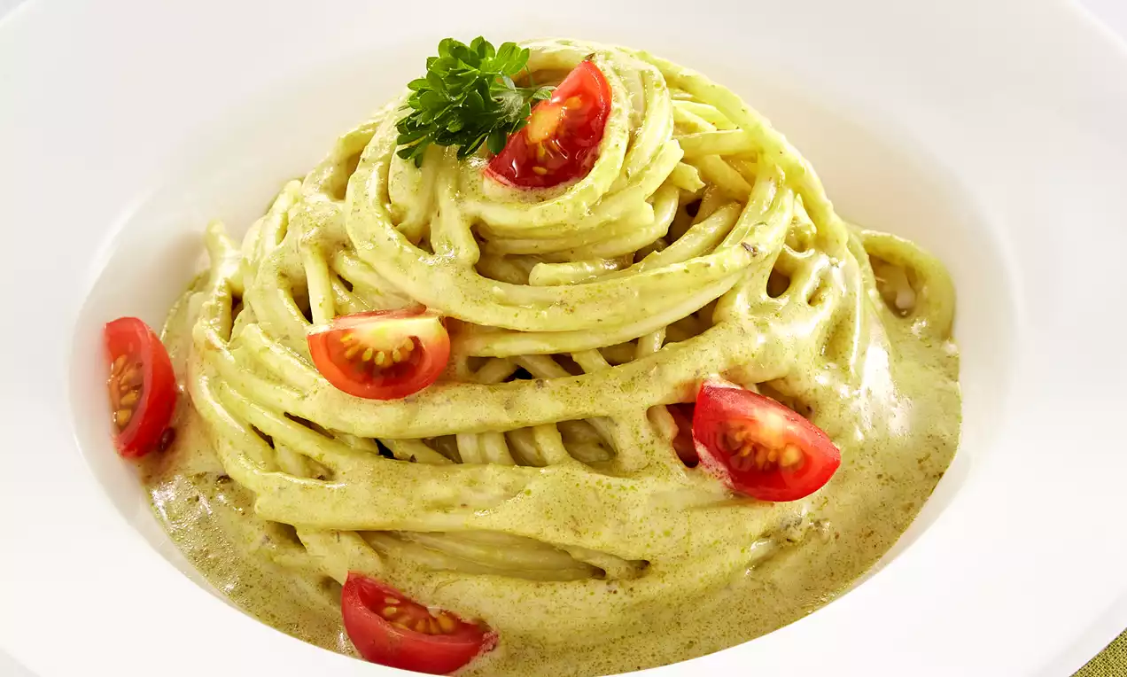 Twisted spaghetti coated in creamy green pesto, garnished with halved red cherry tomatoes and parsley in a white bowl on a green napkin