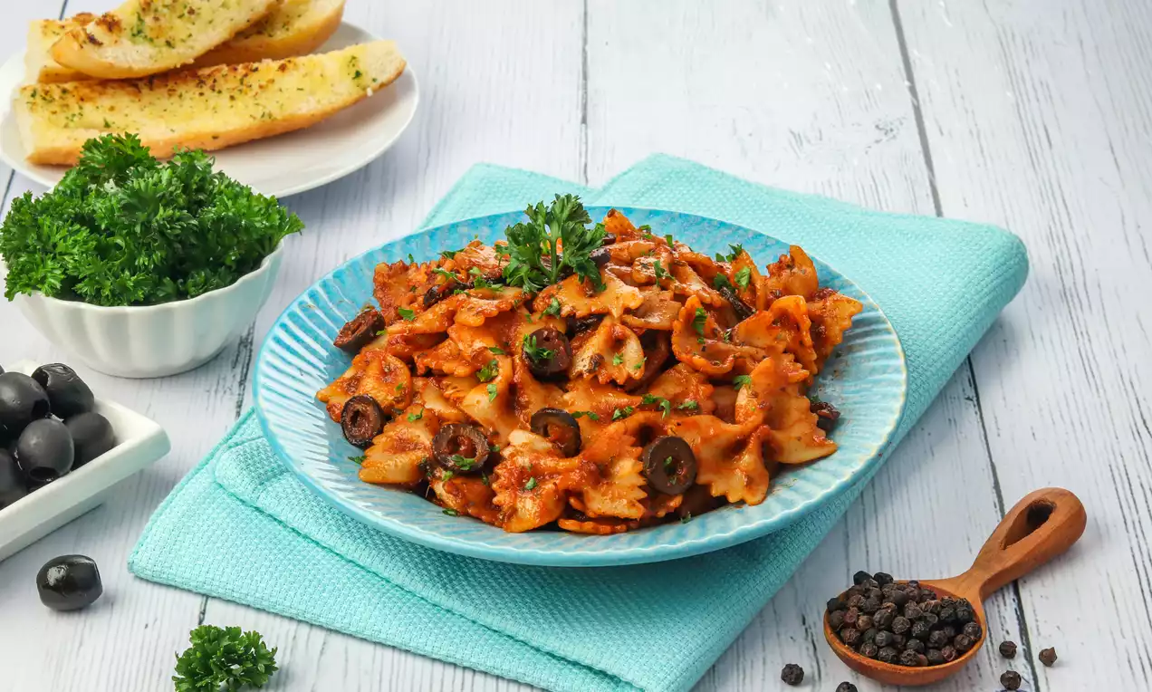 Farfalle pasta coated in rich red sauce with black olive slices and parsley garnish on a blue plate, surrounded by bread and fresh herbs