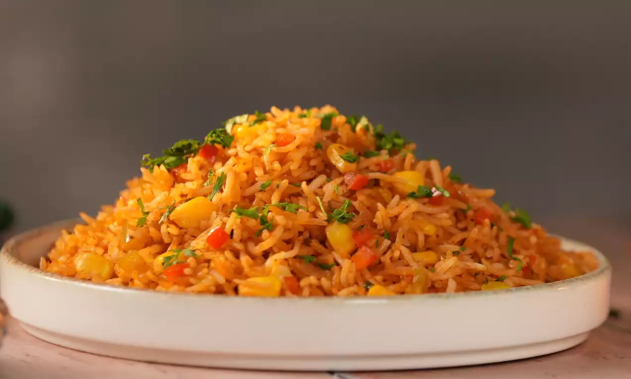 A mound of vibrant orange rice speckled with yellow corn, green herbs and red pepper pieces served on a white plate on a wooden table
